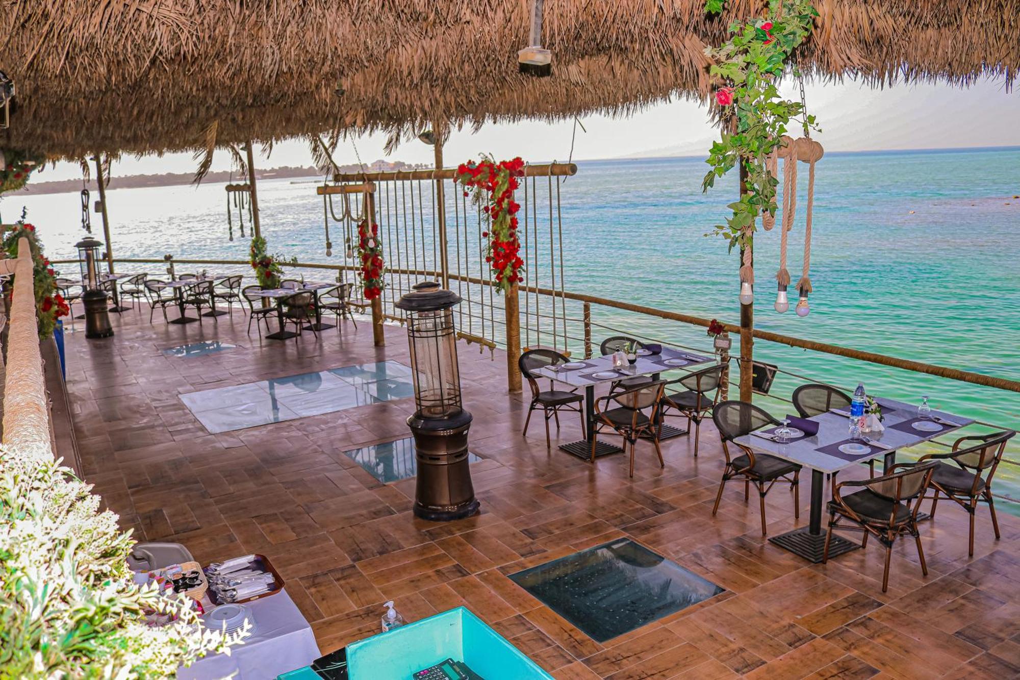 Lagoona Beach Luxury Resort And Spa Manáma Exteriér fotografie The photo depicts a coastal restaurant or dining area with an open-air design. It features a wooden floor and tables set for dining, surrounded by chairs. The space is partially covered by a thatched roof, providing shade while allowing for a view of