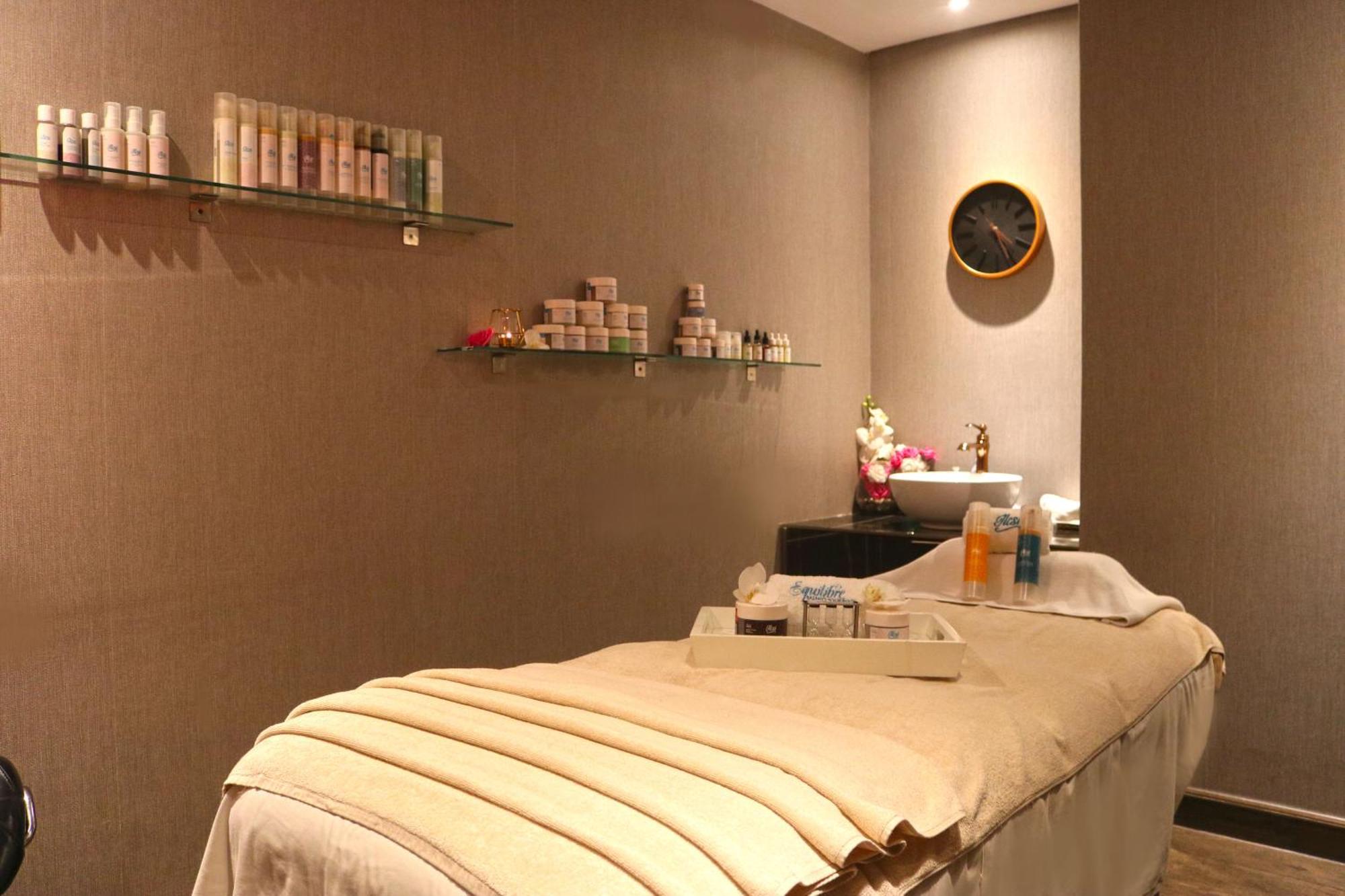 Lagoona Beach Luxury Resort And Spa Manáma Exteriér fotografie The photo shows a spa treatment room. There is a massage table draped with a light-colored blanket, creating a relaxing atmosphere. On the wall, there are shelves displaying various skincare products in bottles and jars. A clock is mounted on the wal