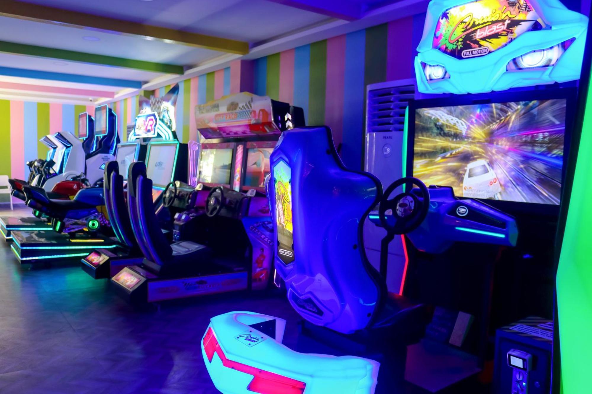 Lagoona Beach Luxury Resort And Spa Manáma Exteriér fotografie The photo shows an arcade area filled with various arcade machines. The setting is illuminated with colorful neon lights, creating a vibrant atmosphere. There are several racing game machines with steering wheels and seats, each accompanied by screen