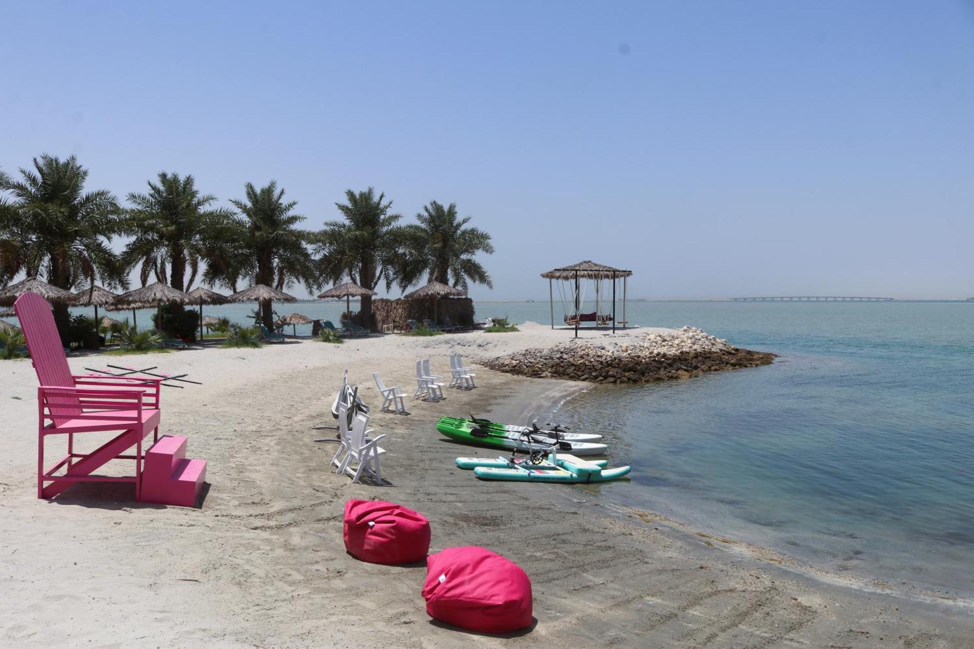 Lagoona Beach Luxury Resort And Spa Manáma Exteriér fotografie The photo depicts a serene beach scene with clear blue water and a sunny sky. In the foreground, there are colorful pink bean bags and a large pink chair on the sand. A few white chairs and loungers are positioned near the water's edge. To the side, 