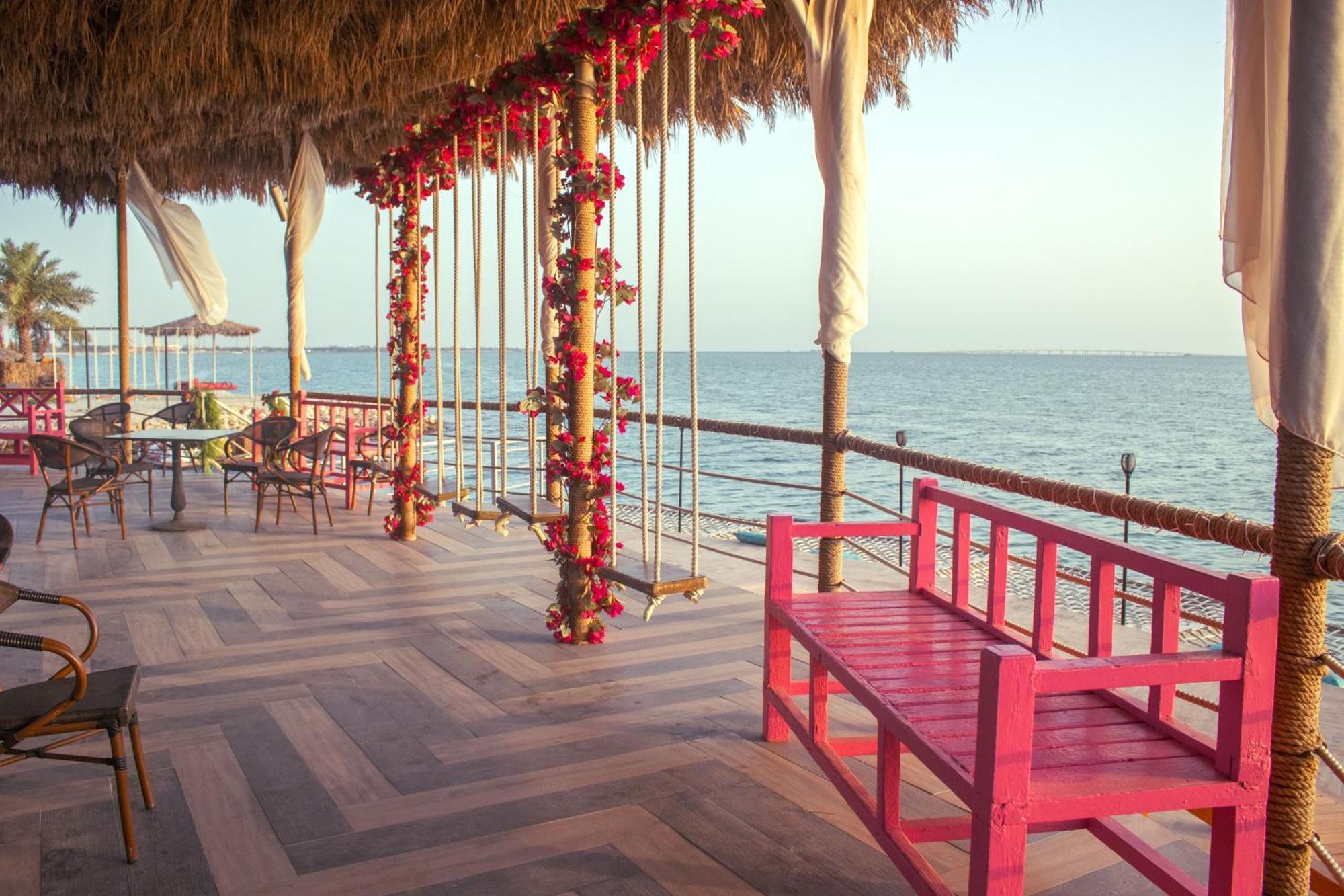 Lagoona Beach Luxury Resort And Spa Manáma Exteriér fotografie The photo depicts a scenic outdoor area by the water, with a wooden deck and a thatched roof. Two swings adorned with flowers are hanging from the ceiling, providing a charming touch to the space. In the foreground, there is a bright pink bench, addi