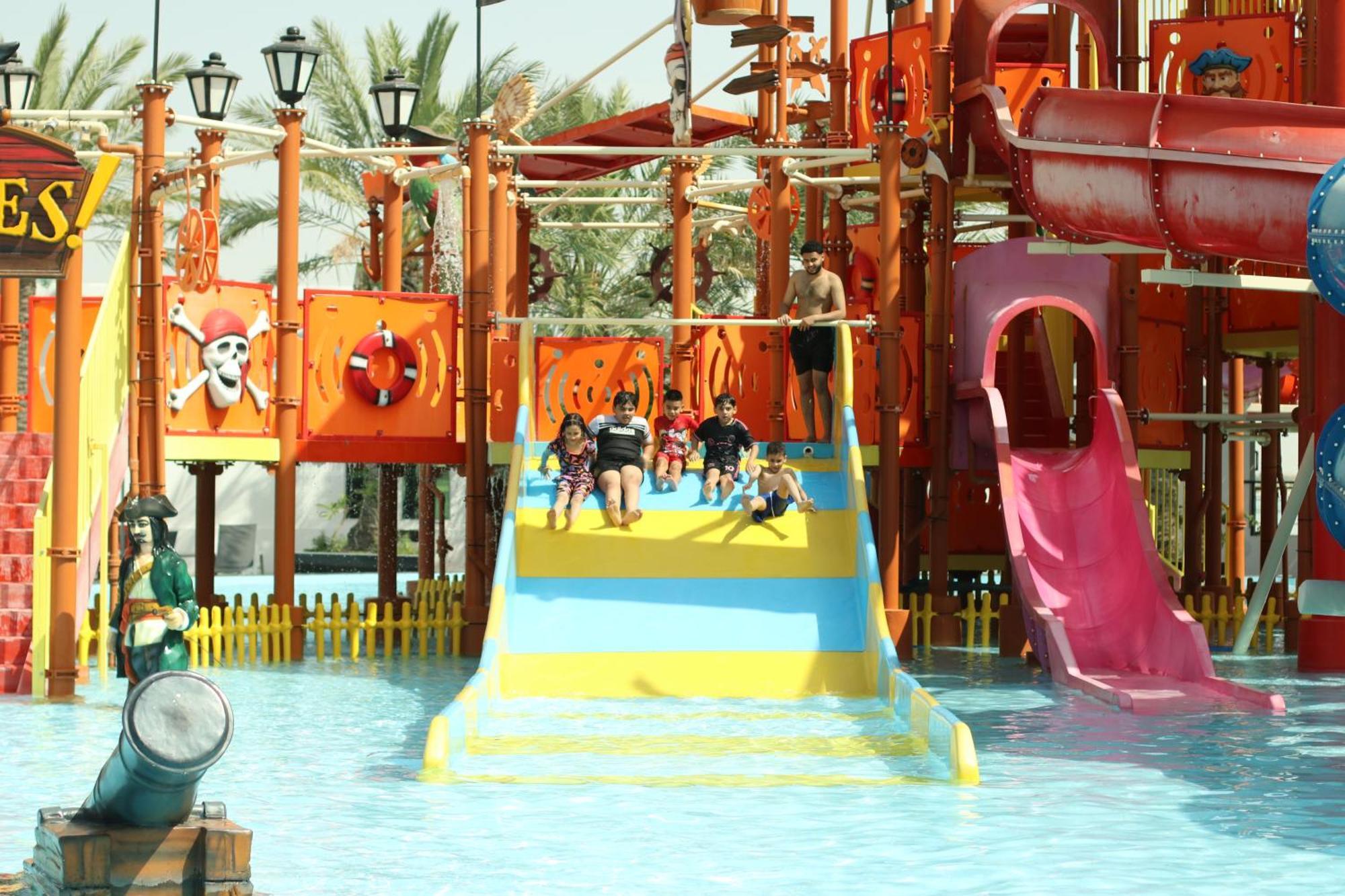 Lagoona Beach Luxury Resort And Spa Manáma Exteriér fotografie The photo shows a colorful water park scene featuring a play area with multiple slides. There are children enjoying themselves, sliding down bright yellow and pink slides into a pool of water. The structure has pirate-themed decorations, including a 