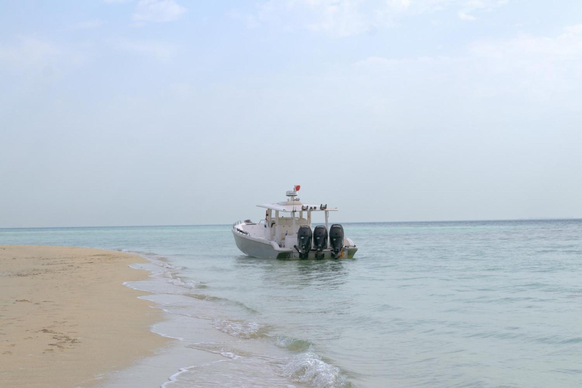 Lagoona Beach Luxury Resort And Spa Manáma Exteriér fotografie The photo depicts a small boat anchored near a sandy beach, partially submerged in calm, clear water. The boat is white and features three outboard motors on its stern. In the background, the sea stretches out toward the horizon under a light, cloudy