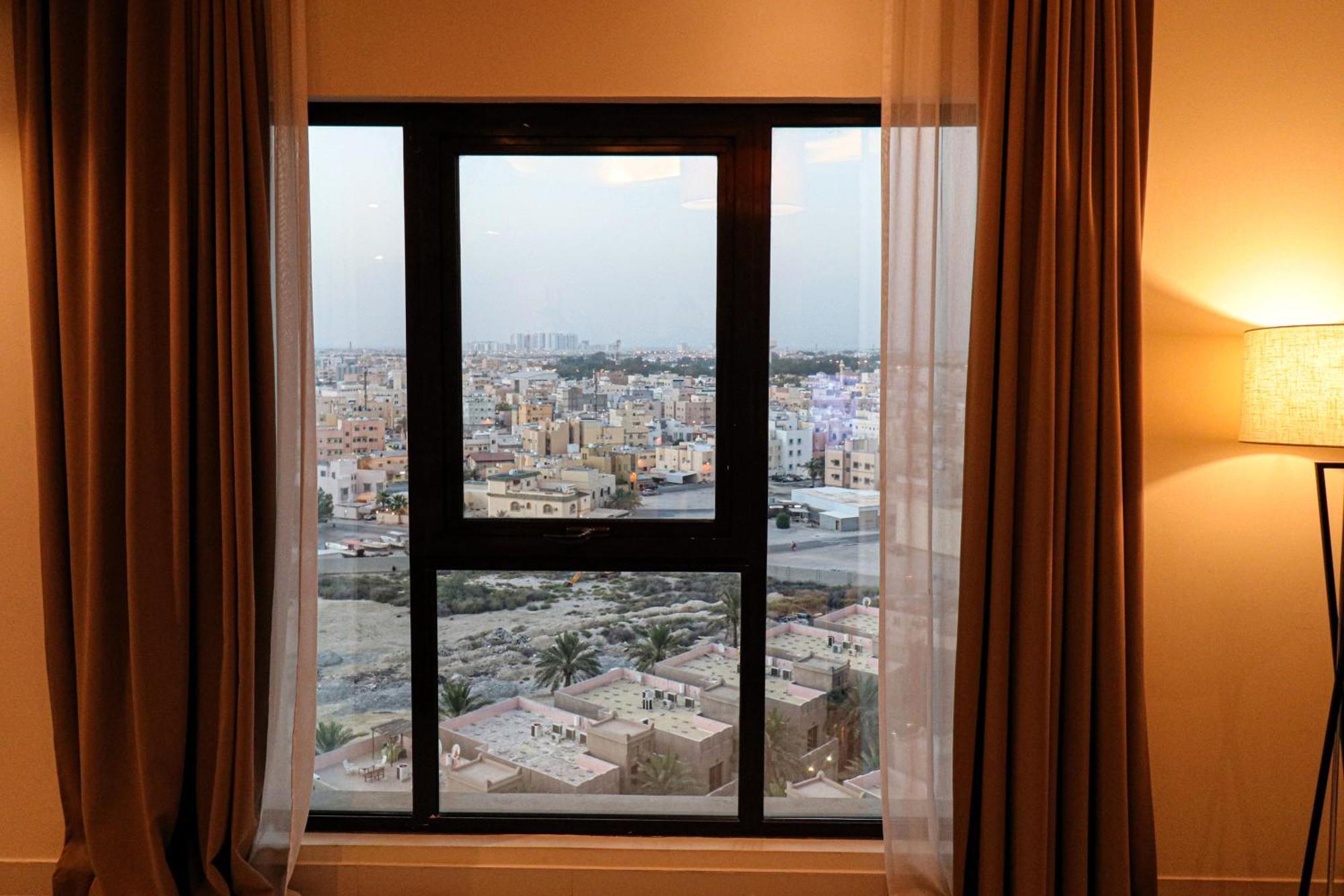 Lagoona Beach Luxury Resort And Spa Manáma Exteriér fotografie The photo shows a window with a black frame and sheer curtains. Through the window, there is a view of a cityscape, displaying a densely populated area with various buildings and rooftops. The sky appears slightly overcast, suggesting a muted lightin