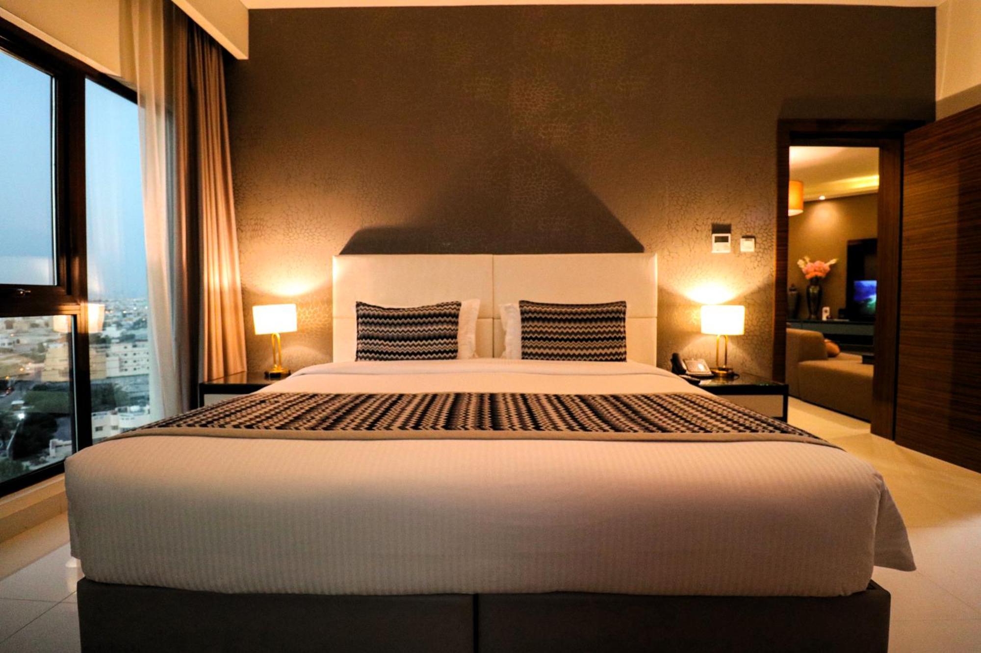 Lagoona Beach Luxury Resort And Spa Manáma Exteriér fotografie The photo depicts a modern hotel room featuring a large bed with a white comforter and two decorative pillows. The headboard is light-colored, and the walls are painted a dark hue. On either side of the bed, there are bedside tables with lamps. The r