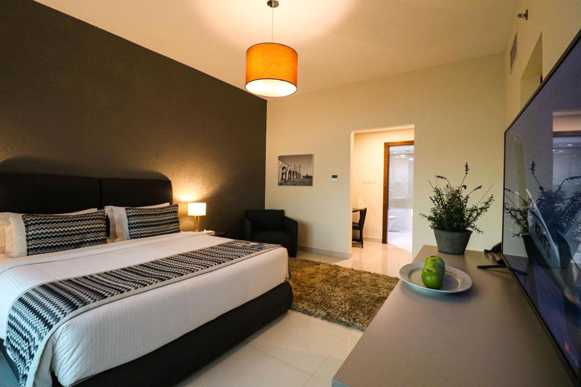 Lagoona Beach Luxury Resort And Spa Manáma Exteriér fotografie The photo shows a modern hotel room. It features a large bed with a neatly made white and dark-colored duvet. On either side of the bed, there are bedside tables with lamps. The wall behind the bed is painted in a dark color, creating a contrast with