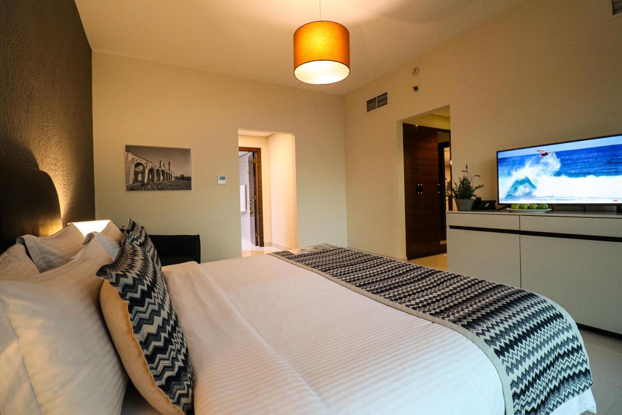 Lagoona Beach Luxury Resort And Spa Manáma Exteriér fotografie The photo shows a well-decorated hotel room. There is a large bed with white linens and decorative pillows featuring a zigzag pattern. On the wall beside the bed is a piece of artwork, and there is a modern television on a white furniture unit opposi