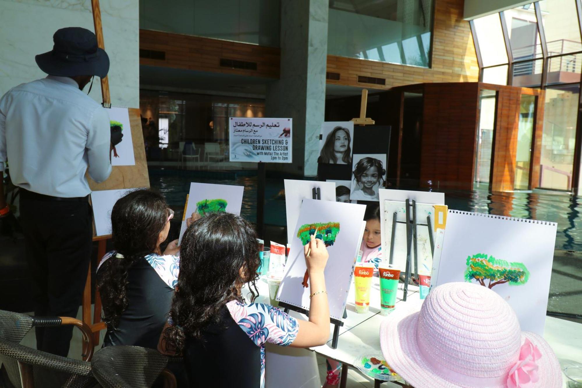 Lagoona Beach Luxury Resort And Spa Manáma Exteriér fotografie The photo shows a group of children and an adult engaged in an art activity near a pool area. The children are seated at a table, painting on canvases or paper, with a focus on trees, as indicated by the green paint they are using. An adult, wearing 