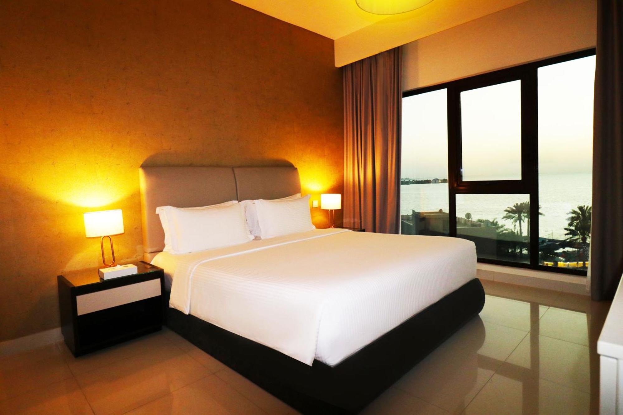 Lagoona Beach Luxury Resort And Spa Manáma Exteriér fotografie The photo shows a modern hotel bedroom with a large, comfortable bed dressed in white linens. The room features warm lighting, including bedside lamps, creating a cozy atmosphere. There are large windows with a view of the outdoors, possibly overlook