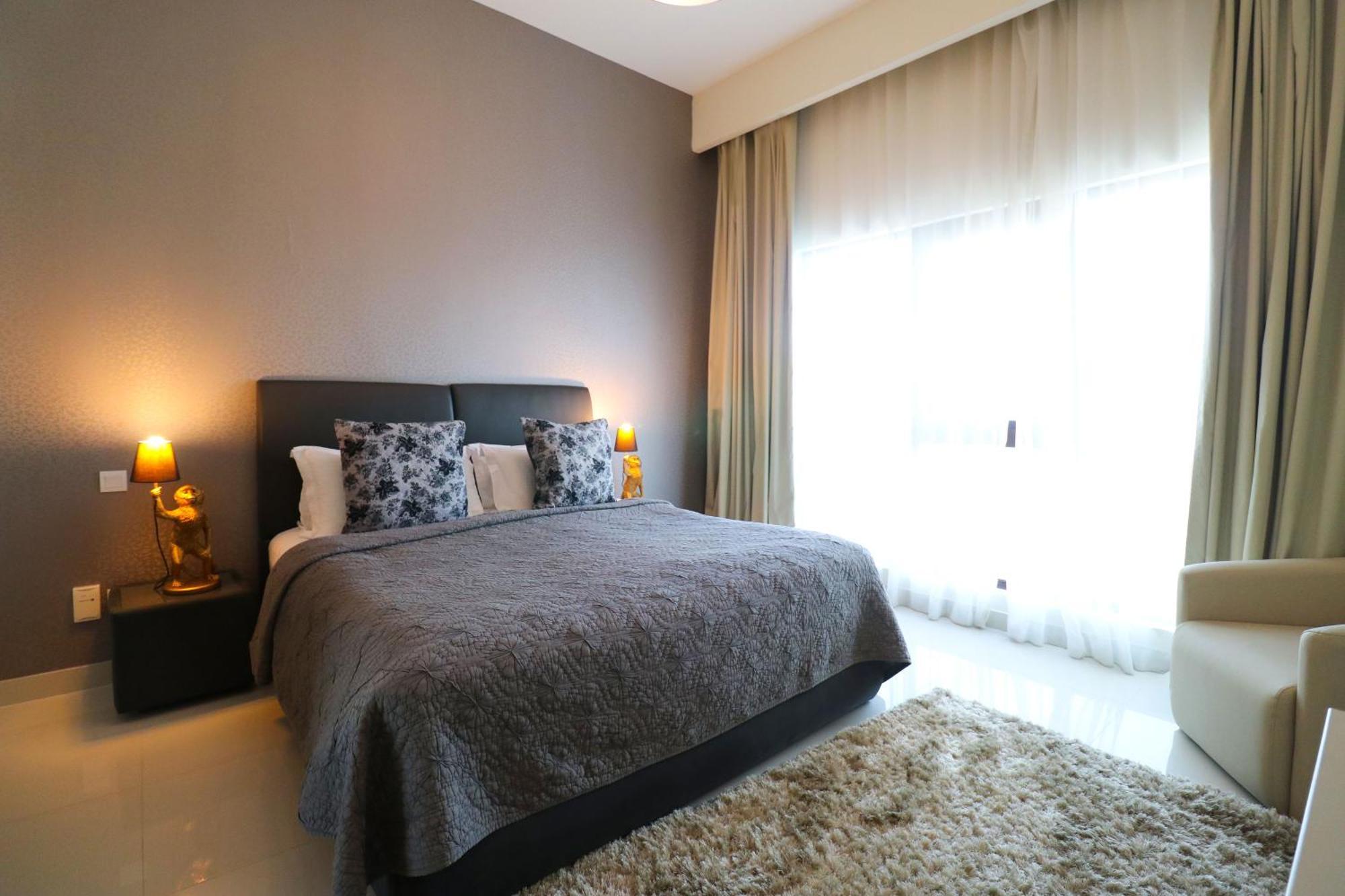 Lagoona Beach Luxury Resort And Spa Manáma Exteriér fotografie The photo shows a stylish bedroom with a modern design. It features a large bed with a dark gray bedspread and decorative pillows. On either side of the bed are two wooden nightstands, each with a lamp. There is a large window covered with light, flo